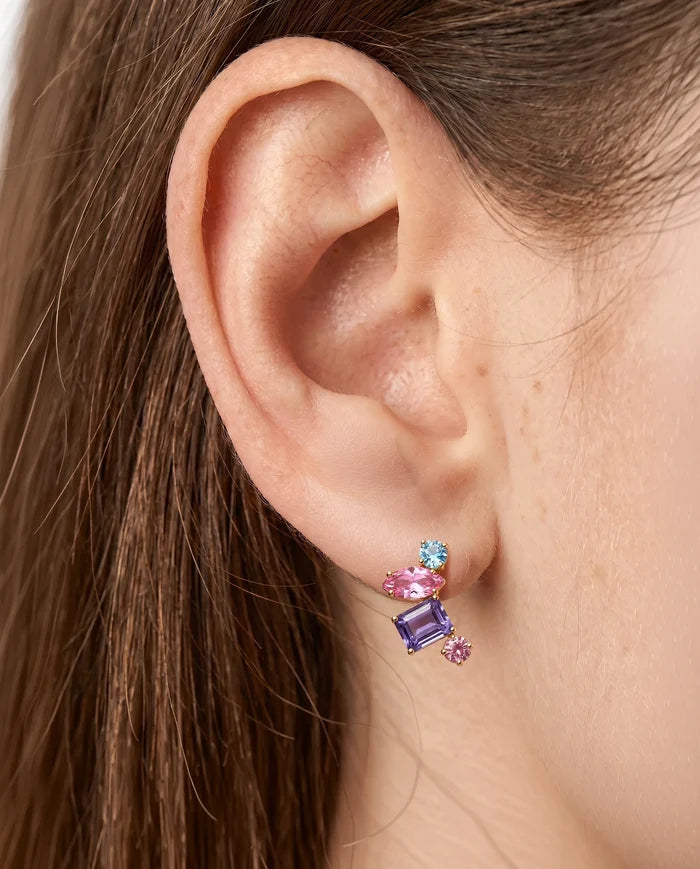 A close-up view of a person wearing a multi-colored gemstone ear climber stud featuring an asymmetrical cluster of light blue, pink, and purple emerald-cut and round-cut stones set on the earlobe.