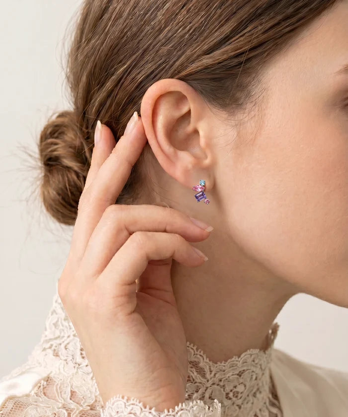 A close-up of a woman's ear wearing a small colorful earring. The earring features a silver-toned twisted wire design with small pink, purple, and blue gemstone-like accents.
