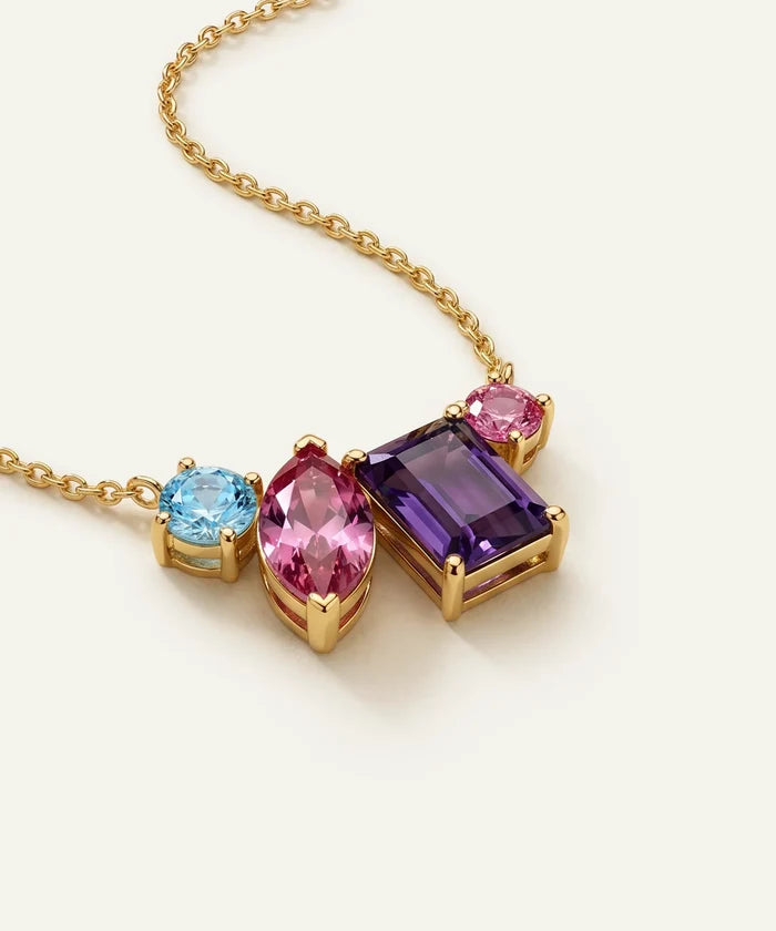 A gold chain necklace featuring a multi-stone pendant with a light blue oval gemstone, a marquise-cut pink gemstone, and a rectangular purple gemstone, all set in gold on a light cream background.
