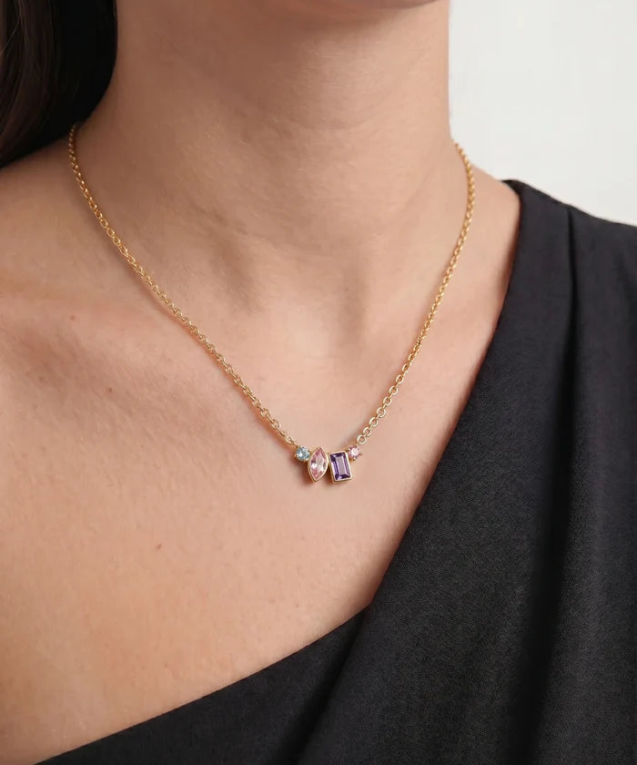 A close-up of a model wearing a delicate gold chain necklace with a central cluster of small beads in lavender, pink, and light blue, paired with a black top.