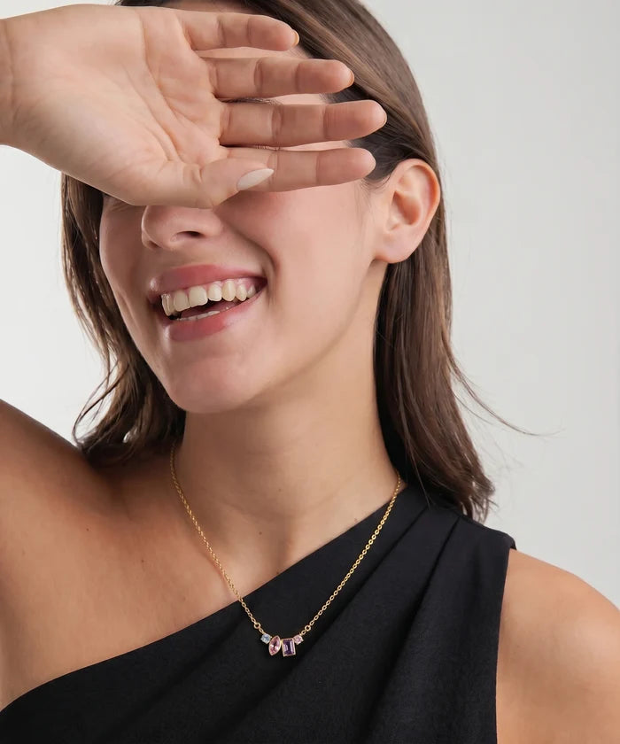 A close-up lifestyle shot of a woman wearing a delicate gold chain necklace with a bow-shaped pendant set with pink gemstones. She is smiling and covering one eye with her hand, wearing a black one-shoulder top against a light gray background.