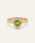 A heart-shaped peridot ring in a gold setting, surrounded by a halo of small clear stones and a pavé band, shown on a light cream background.