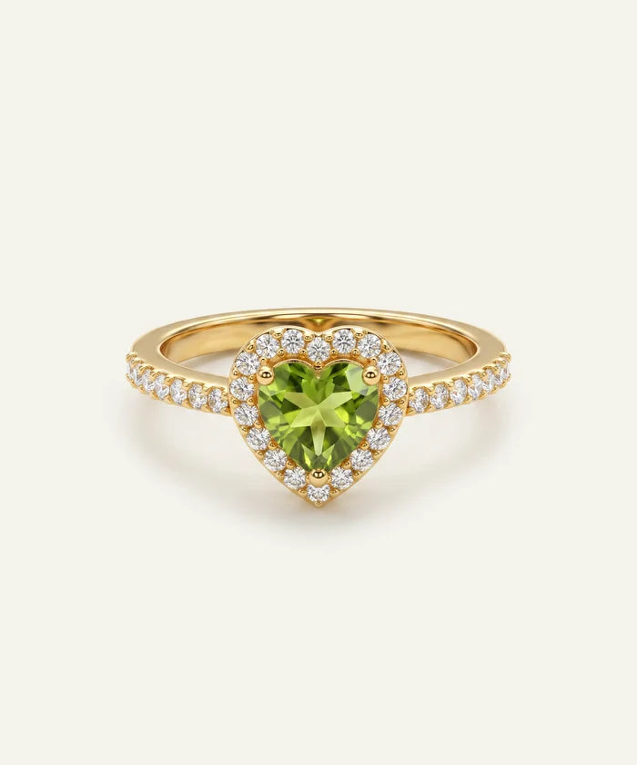 A heart-shaped peridot ring in a gold setting, surrounded by a halo of small clear stones and a pavé band, shown on a light cream background.