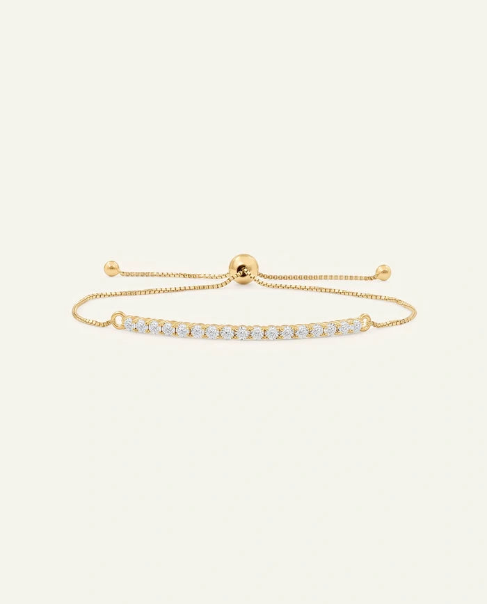 A delicate gold-tone adjustable bracelet featuring a curved bar set with small clear stones, displayed on a light cream background.