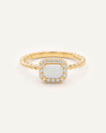 A gold ring featuring a square-shaped white opal center stone surrounded by a halo of small clear stones, with a twisted gold band, shown on a light off-white background.