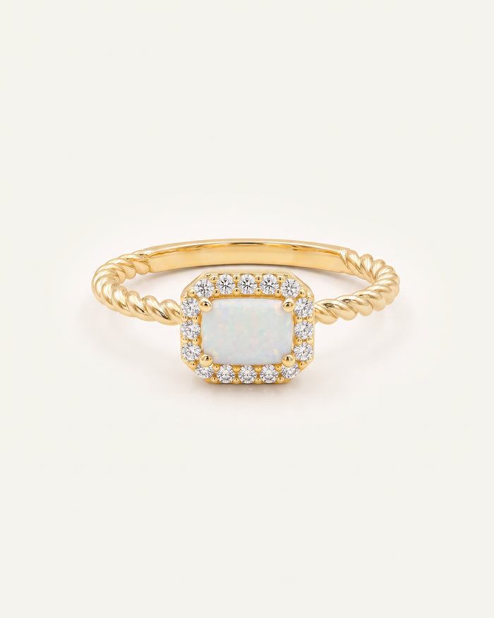 A gold ring featuring a square-shaped white opal center stone surrounded by a halo of small clear stones, with a twisted gold band, shown on a light off-white background.