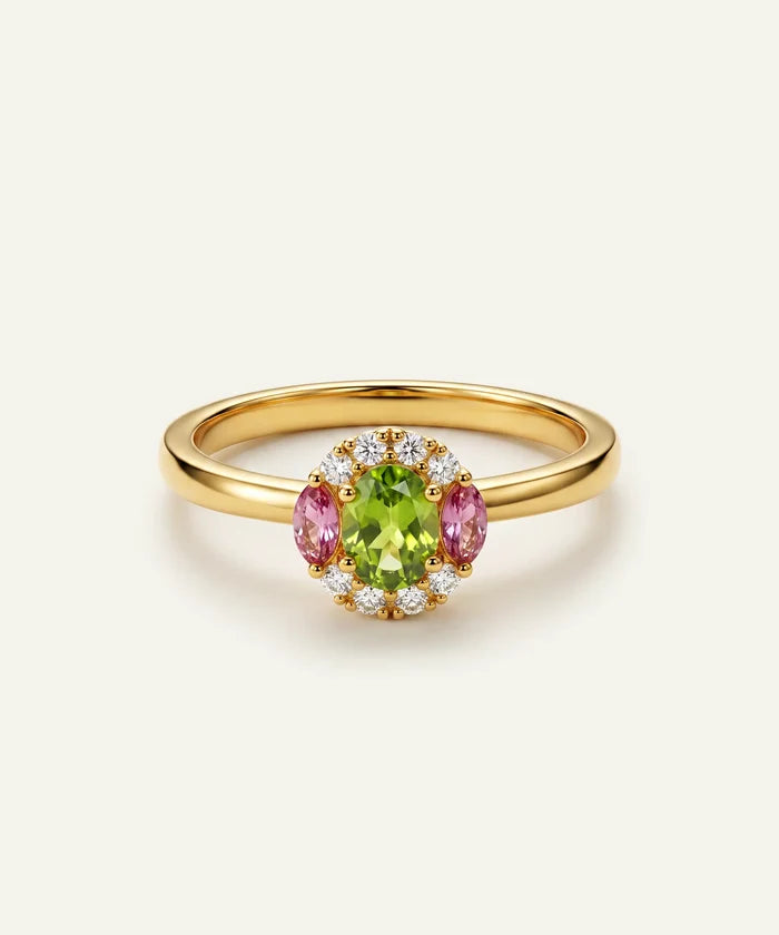 A yellow gold ring featuring a central oval green peridot gemstone surrounded by a halo of small clear stones and two marquise-cut pink sapphires, set against a light cream background.