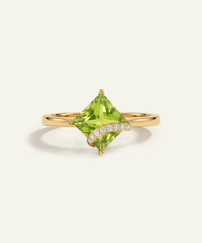 A yellow gold ring featuring a large, faceted green gemstone in a diamond-cut shape, accented by a curved row of small clear stones on the band, set against a light cream background.