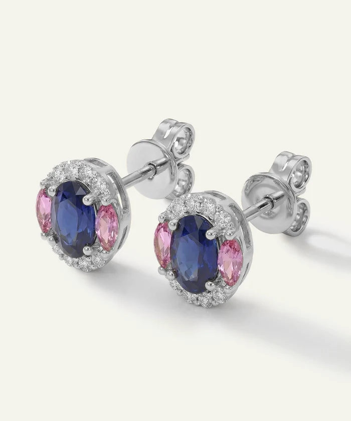 A pair of oval stud earrings with deep blue oval center stones, surrounded by a halo of small clear stones and accented by pink marquise-cut side stones, set in a silver-toned metal on a light cream background.