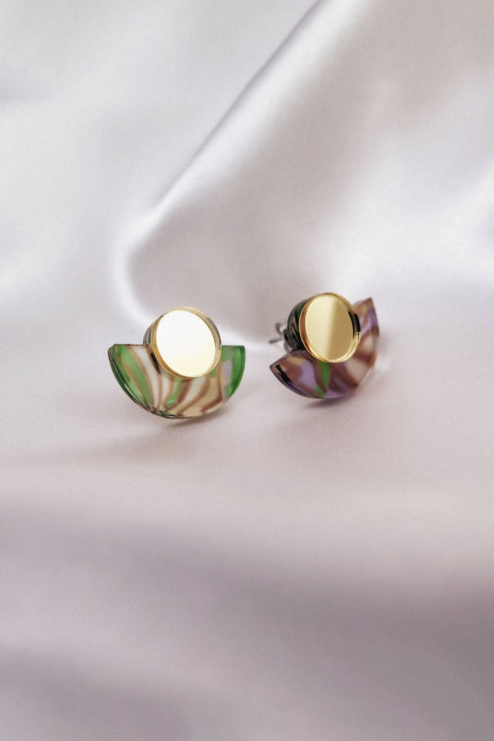 A pair of minimalist geometric stud earrings featuring round gold studs and semicircular marbled green and purple acrylic accents, displayed on soft, white fabric.
