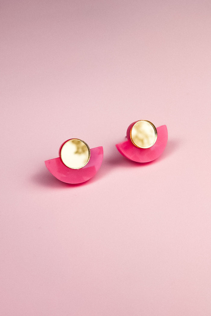 A pair of minimalist geometric stud earrings with round, gold-edged, marble-like white studs and bright pink, semicircular bases, displayed on a soft pink background. Hypoallergenic and lightweight for comfortable wear.