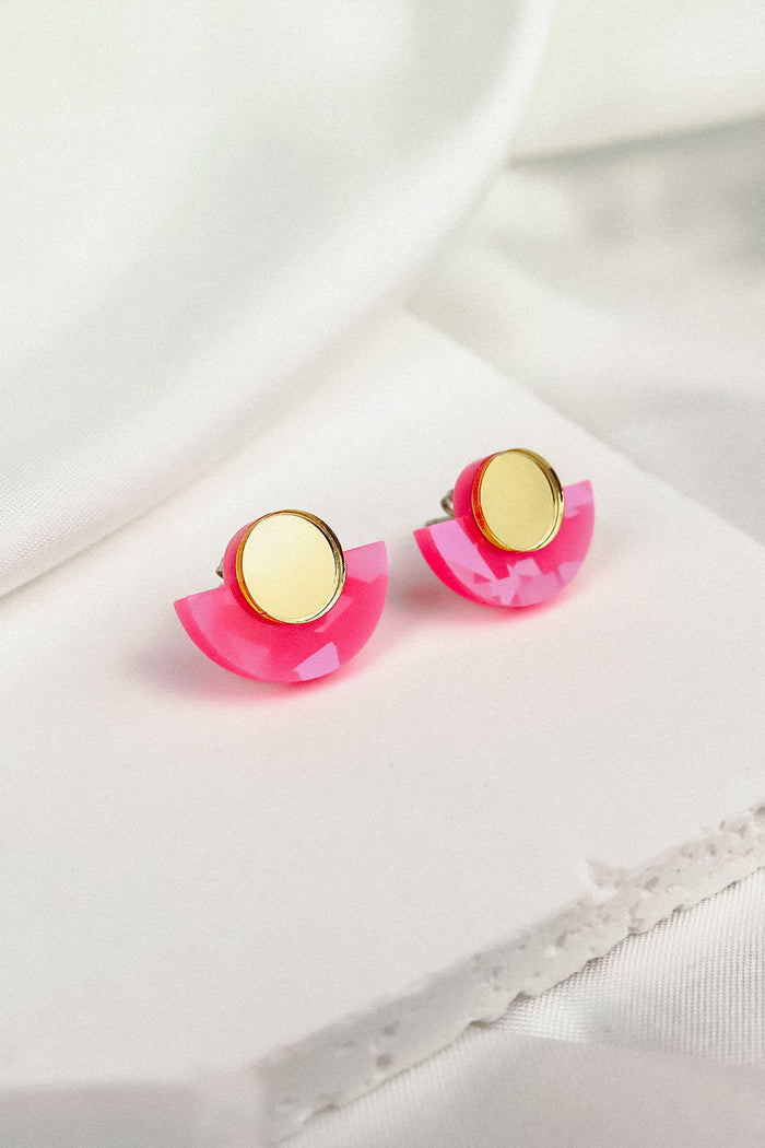 Minimalist Geometric Stud Earrings featuring gold circular tops and pink acrylic semicircles. These lightweight, hypoallergenic studs are displayed on a white fabric surface.