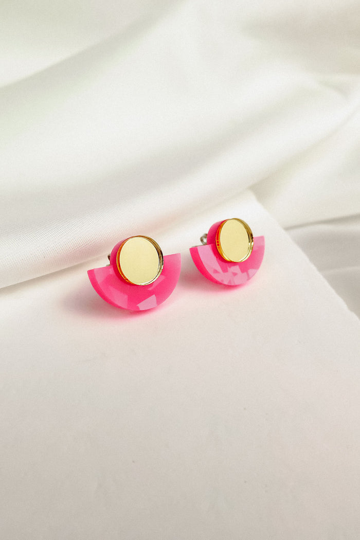 Minimalist Geometric Stud Earrings featuring gold circular tops and bright pink, semi-circle bases are displayed on soft white fabric. These lightweight, hypoallergenic studs add a playful touch to any look.