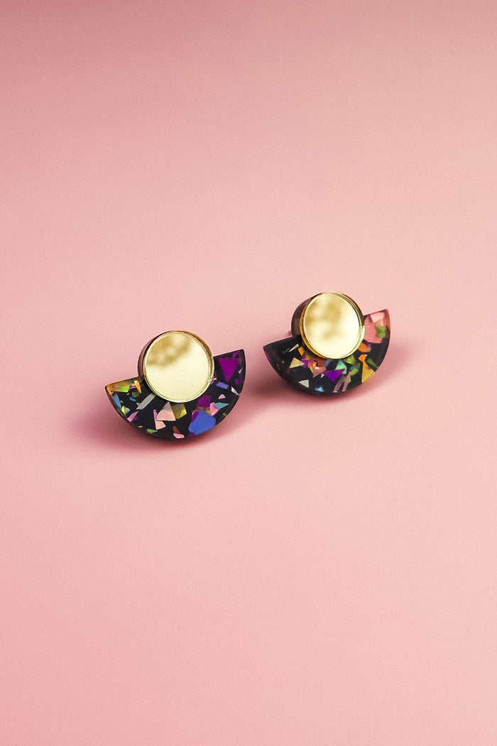 A pair of minimalist geometric stud earrings with gold circular tops and black semicircular bases featuring vibrant, multicolored confetti-like patterns, these hypoallergenic, lightweight pieces are displayed on a pink background.