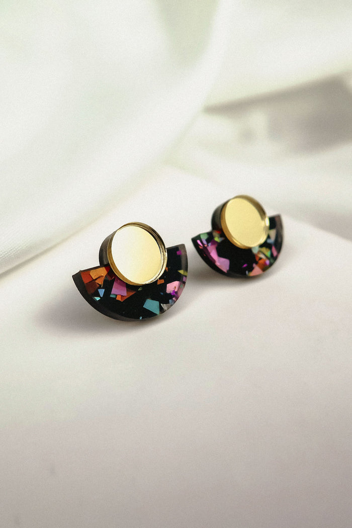 Minimalist Geometric Stud Earrings featuring round gold tops and semicircular black bases with colorful patterns. These lightweight, hypoallergenic earrings rest on a white surface with soft, light fabric in the background.