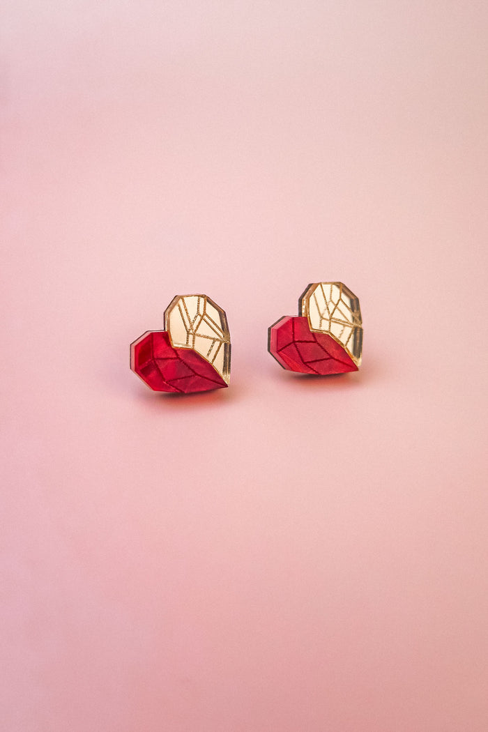Two origami hearts, each half red and half gold, are placed side by side on a smooth, light pink background, resembling handcrafted earrings or romantic jewelry.