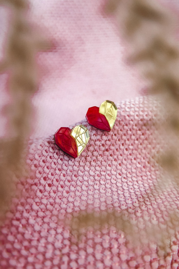 A pair of handcrafted red and gold geometric heart-shaped earrings rest on a textured pink fabric, with blurry plant stems in the foreground—perfect romantic jewelry for any occasion.
