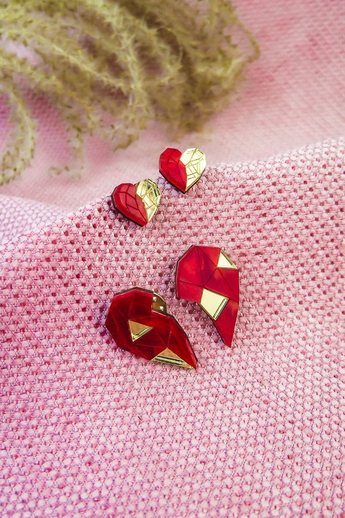 Red and gold geometric heart studs and pins, perfect as romantic jewelry, are displayed on textured pink fabric, with decorative plant stems in the background.