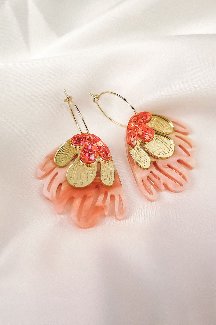 A pair of Organic Floral Hoops featuring gold, petal-shaped pieces and pink, coral-colored acrylic flowers, arranged in a floral design, displayed on a soft white fabric background.