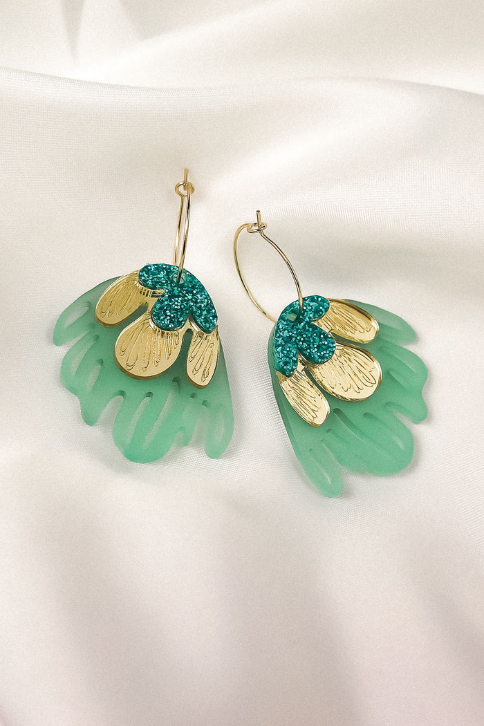 A pair of handcrafted earrings with gold metallic petals, glittery green accents, and translucent green acrylic shapes resembling leaves, displayed on white satin fabric—perfect for adding a touch of tropical elegance.