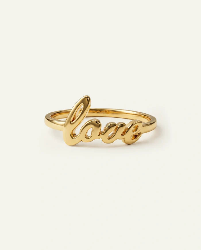 A gold script ring spelling 'love' in cursive, shown on a plain off-white background.
