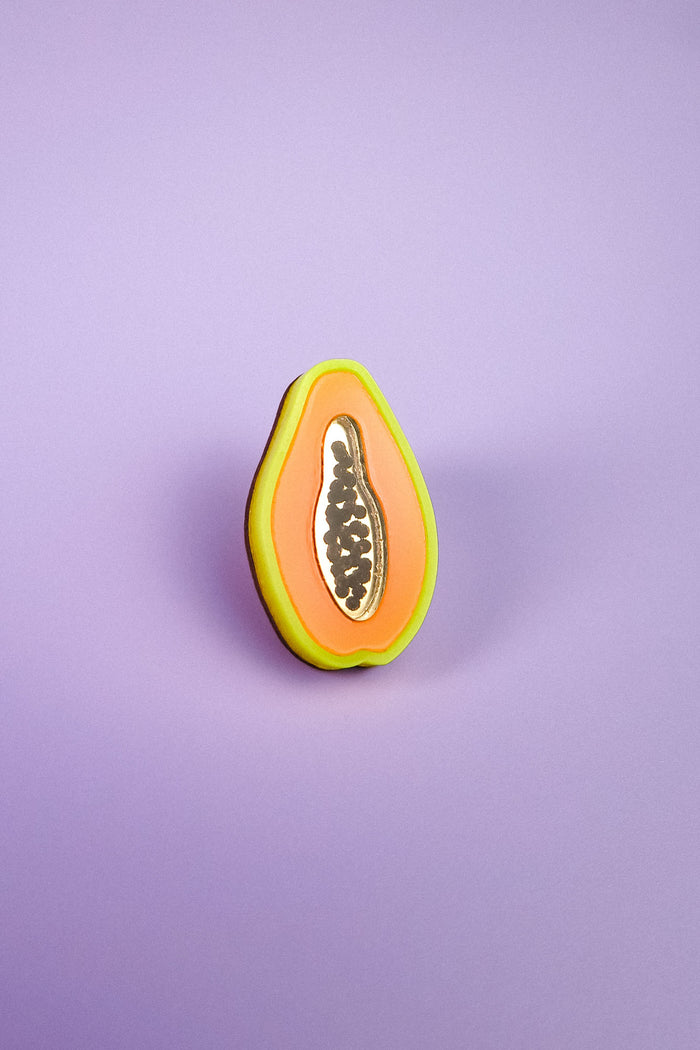 A handcrafted cookie decorated to look like a sliced papaya, with neon orange, yellow, and green icing plus black seeds, stands upright on a purple surface with a purple background—just like a Papaya Pin come to life.