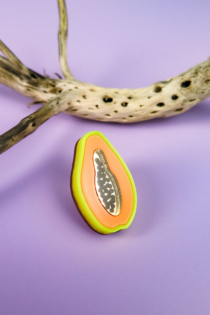 A handcrafted, colorful papaya-shaped cookie with icing details sits on a purple surface next to a dried, spotted branch, reminiscent of a neon Papaya Pin.