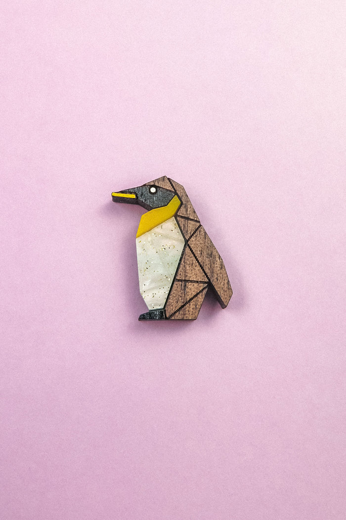 A wooden, geometric Penguin Pin with yellow, white, and black accents serves as a charming accessory, set against a light pink background.