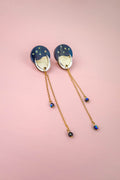 A pair of earrings with blue circular tops reminiscent of the night sky, gold accents, and long gold chains with small blue beads, displayed on a pink background.