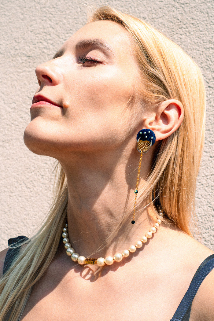 A woman with long blonde hair wears a pearl necklace and a bold blue and gold earring with chain detail, evoking festival magic as she poses in sunlight against a textured light wall.