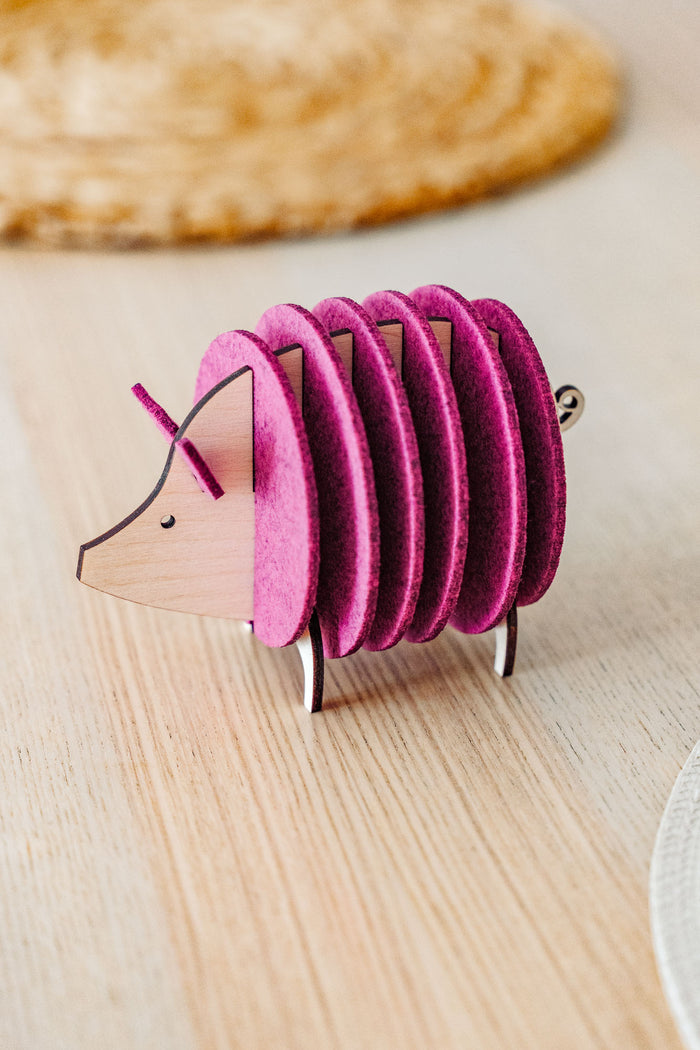 A small, decorative, pink and wood-colored armadillo figurine with a segmented, accordion-like body stands on a light wooden surface—perfect as a quirky housewarming gift.