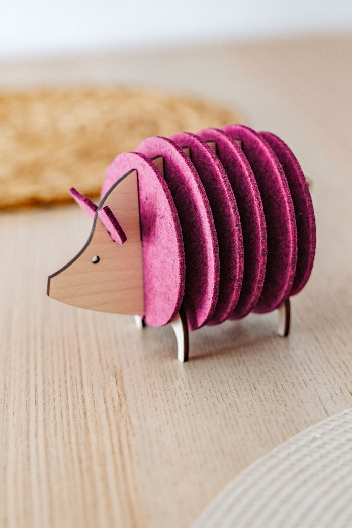 A wooden and felt coaster holder shaped like a hedgehog, with pink felt discs forming the body, sits on a light wooden table—a charming housewarming gift for any home.