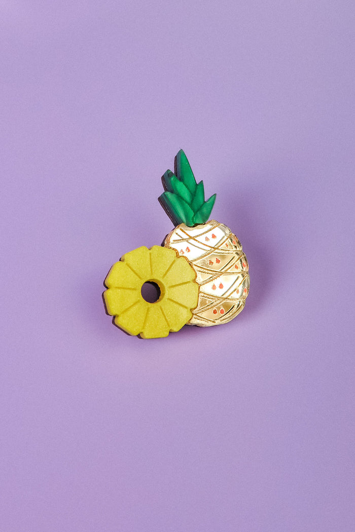 A handcrafted Pineapple Pin with green leaves and gold detailing stands on a light purple background, next to a yellow ring-shaped accessory resembling a sliced pineapple.