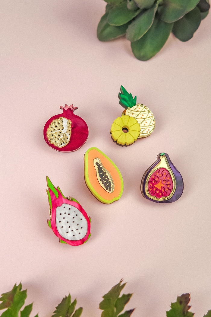 Five colorful fruit-shaped enamel pins—pomegranate, pineapple, papaya, fig, and dragon fruit—are arranged on a light pink surface with green succulent plants at the edges, making a vibrant handcrafted fashion accessory set.
