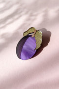 A decorative brooch shaped like a plum with two green leaves, crafted from acrylic and recycled wood, rests on a pale pink surface with soft shadows.