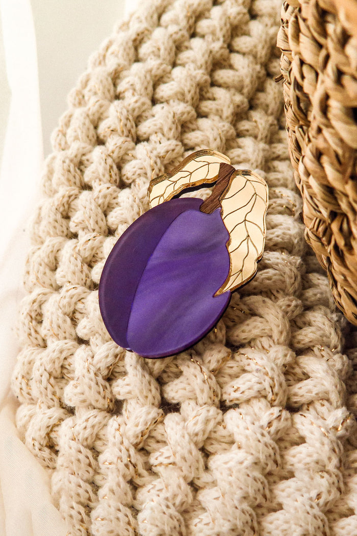 A purple enamel eggplant brooch with gold-outlined leaves rests on a chunky, cream-colored knit blanket beside a woven basket—crafted by a family business.