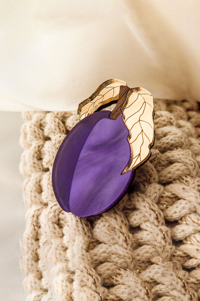 A handmade purple plum-shaped brooch with two gold and ivory leaves is attached to a chunky beige knit fabric. Cream-colored fabric appears in the background.