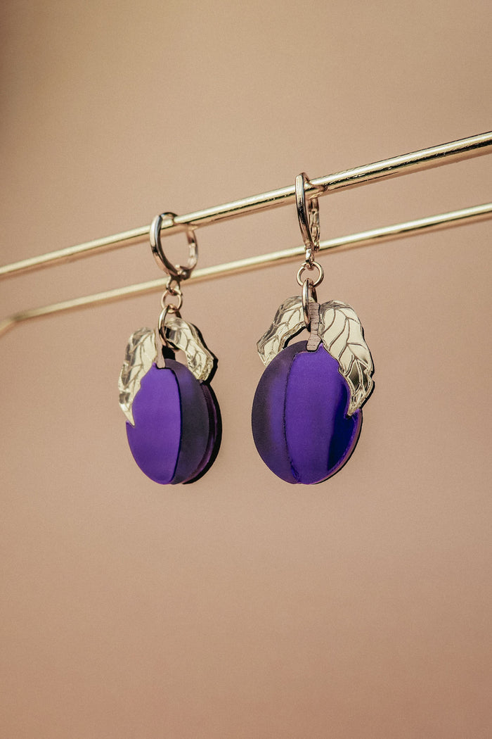 A pair of elegant purple plum earrings with silver leaf accents hang from a gold-colored rod against a beige background.
