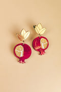 A pair of handcrafted earrings shaped like pomegranates with gold leaves on top and intricate white and gold seed patterns on a red background, displayed on a beige surface, showcasing expert craftsmanship.
