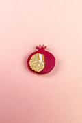 A handcrafted red pomegranate ornament with a gold, textured section showing seeds sits on a light pink background—an elegant fashion accessory for any collection.