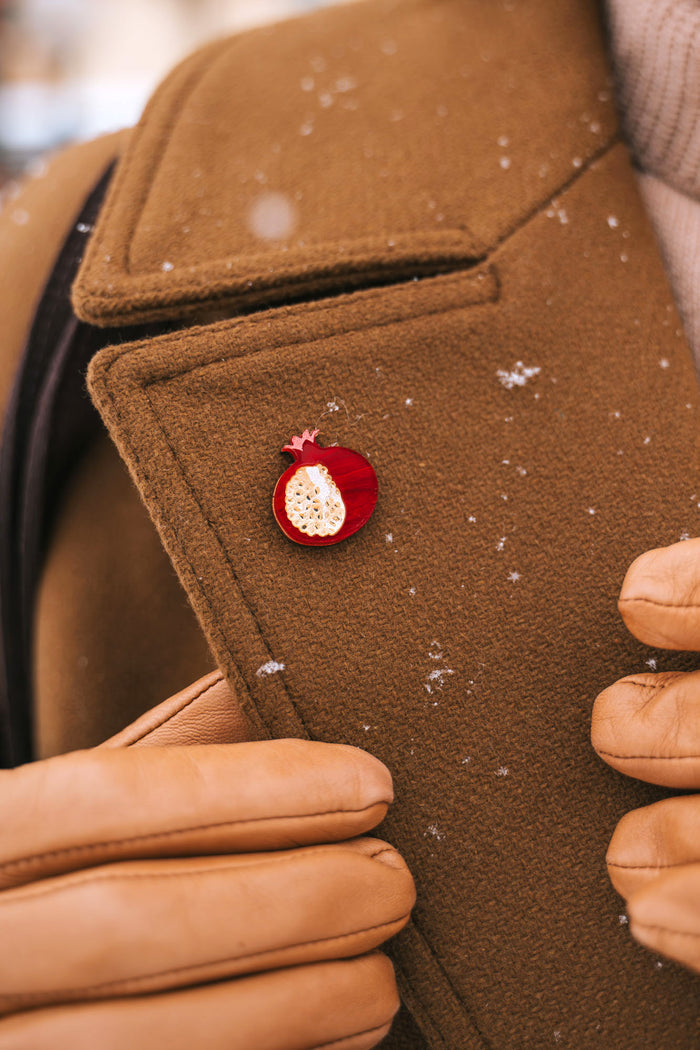 A person wearing a brown coat and tan gloves adjusts the lapel, which is decorated with a handcrafted Pomegranate Pin—a unique fashion accessory. Snowflakes are visible on the coat.