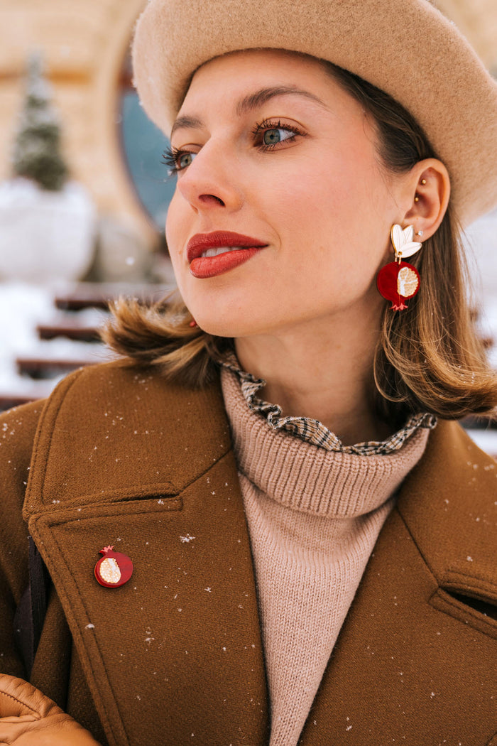 A stylish woman in a beige beret, brown coat, and tan gloves poses outdoors in snowy weather. She wears red and gold handcrafted earrings and a matching Pomegranate Pin fashion accessory, with snowflakes visible on her coat.