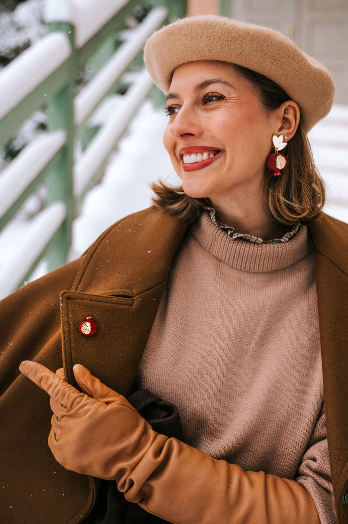 A smiling woman wearing a beige beret, brown coat, tan gloves, and statement earrings with red and gold accents sits outside in a snowy setting, pointing to a handcrafted Pomegranate Pin—a unique fashion accessory on her coat.