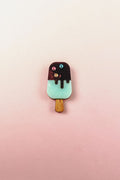 A handcrafted Popsicle Pin shaped like a popsicle with a wooden stick, mint green bottom, chocolate top, and three colored candy decorations, set against a pink and cream gradient background.