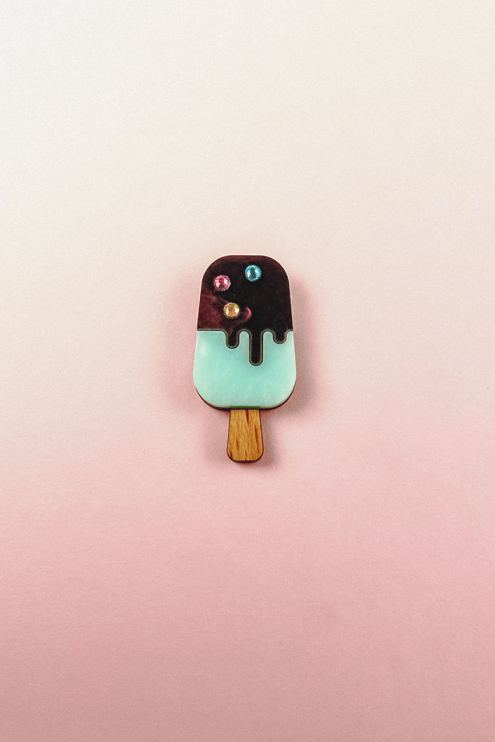 A handcrafted Popsicle Pin shaped like a popsicle with a wooden stick, mint green bottom, chocolate top, and three colored candy decorations, set against a pink and cream gradient background.