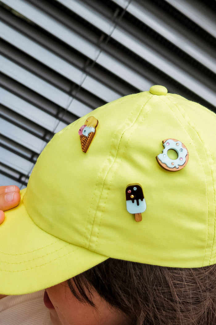A person wears a bright yellow cap adorned with three cute handcrafted pins: an ice cream cone, a donut pin sparkling with rhinestones and sprinkles, and a chocolate-covered popsicle. Venetian blinds are in the background.