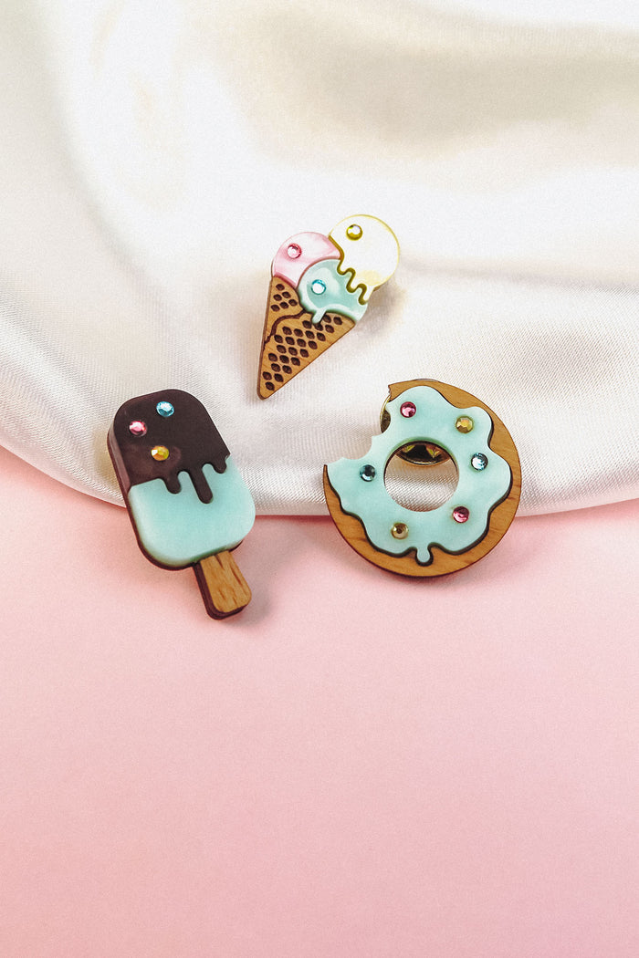 Three wooden pins shaped like desserts—a popsicle, an ice cream cone, and a handcrafted Donut Pin—decorated with pastel icing and colorful sprinkles, are displayed on a pink and white background.