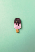 A small, pink and brown handcrafted popsicle pin with glittery gem accents and rhinestones, placed on a mint green background.