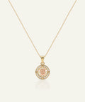 A gold chain necklace with a circular pendant featuring a rose gold center engraved with the number 15, surrounded by a halo of small clear stones, on a light cream background.