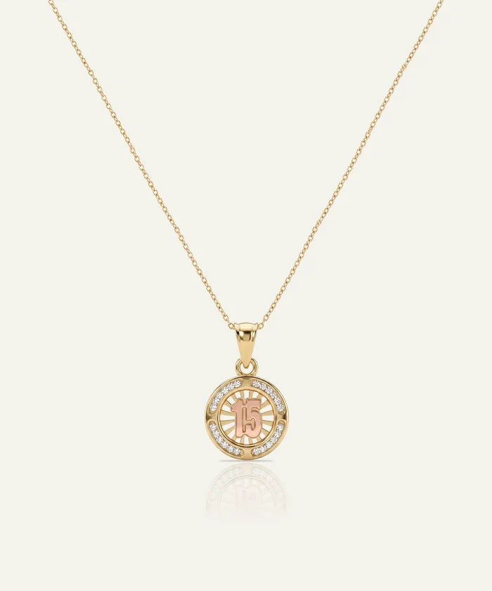 A gold chain necklace with a circular pendant featuring a rose gold center engraved with the number 15, surrounded by a halo of small clear stones, on a light cream background.